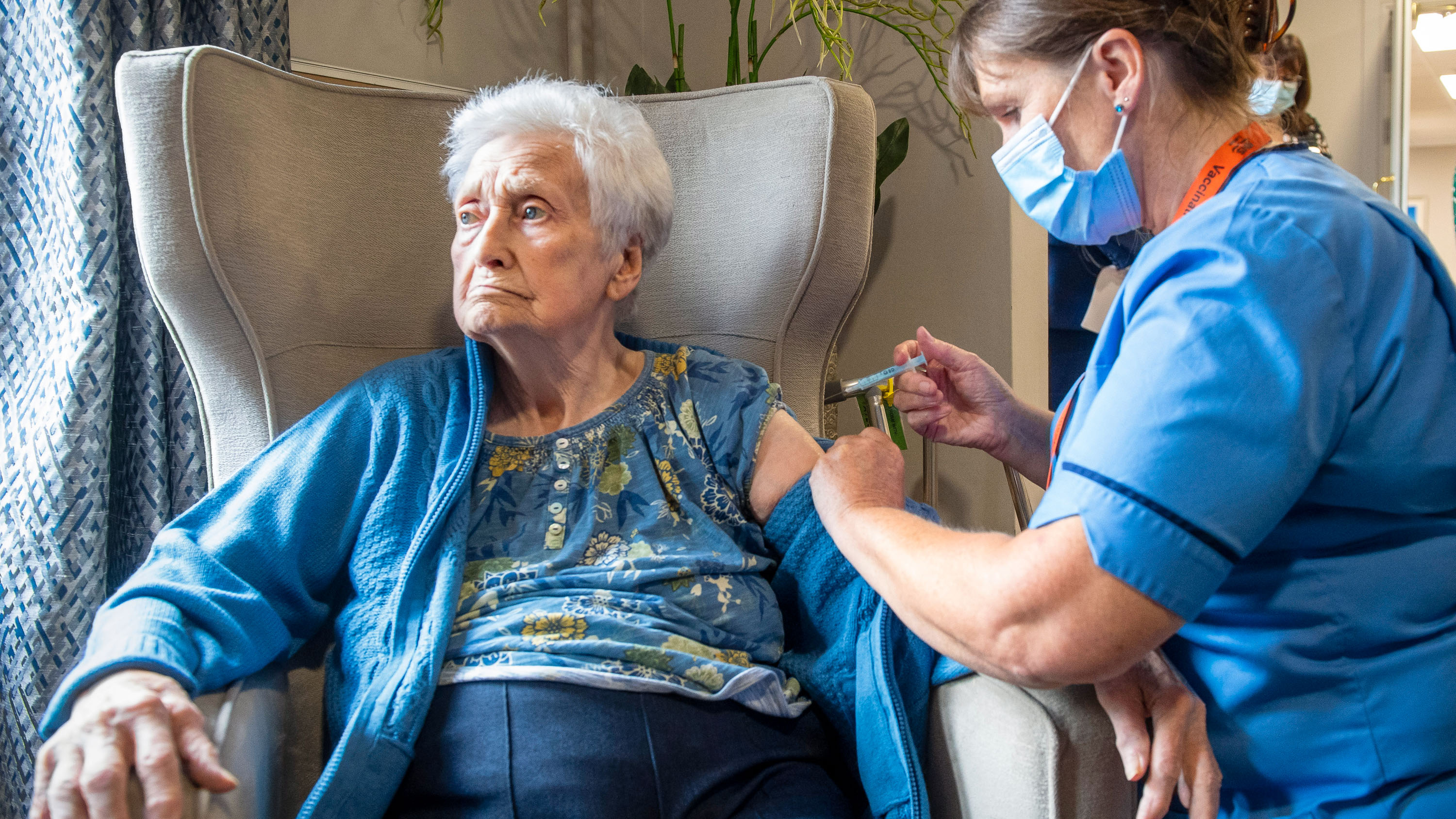 Nurse administering a vaccine booster to an elderly patient seated in an armchair by a window